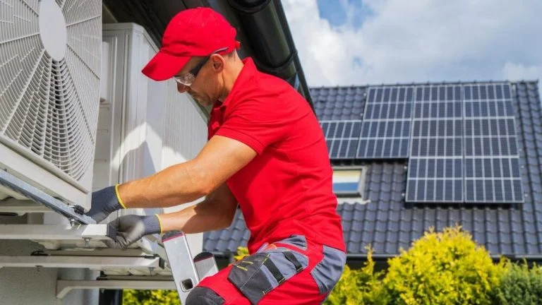 Affordable HVAC services technician in red shirt and cap servicing energy-efficient outdoor unit beside house with solar panels.