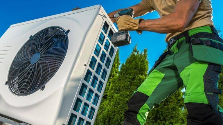 Green and black uniformed technician using a power drill on an exterior component of a high-efficiency HVAC system.